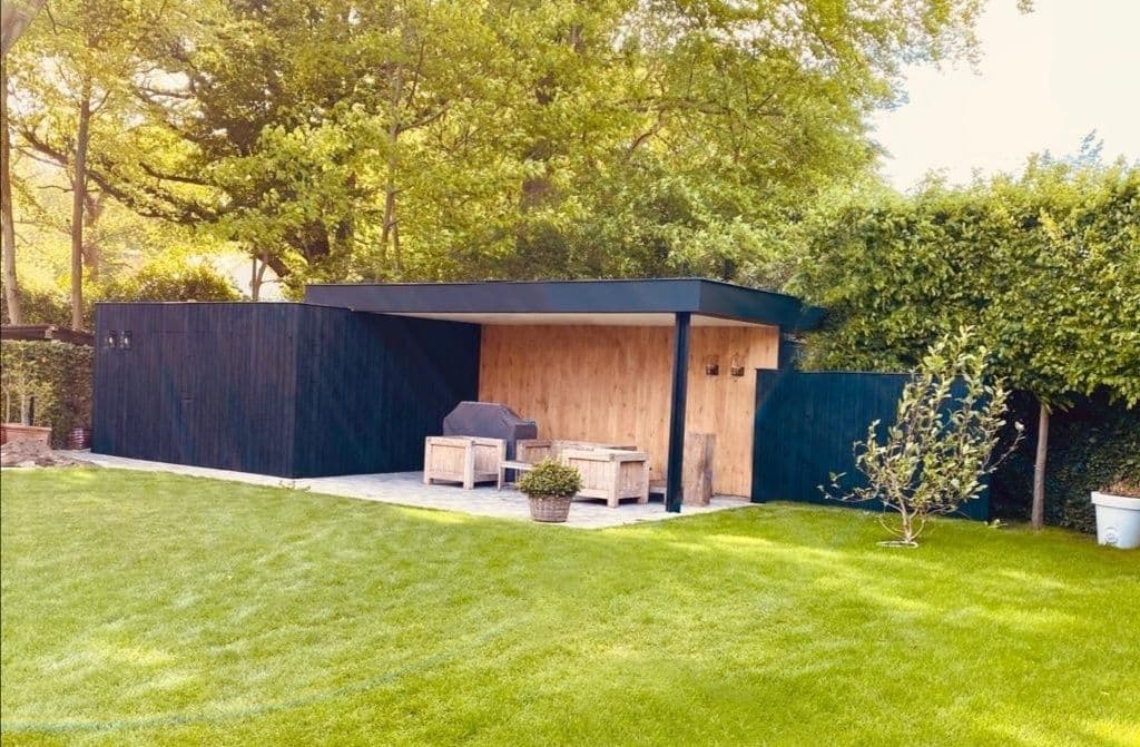 Donkerblauwe tuinopslag met houten paneel onder afdak; zithoek met houten bankjes, groene gazon en bomen op de achtergrond.