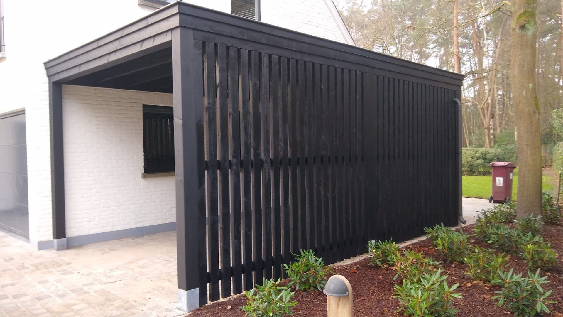Zwarte houten afscheiding langs een witte woning met overkapping. Voorgrond met groene planten en een bruine afvalcontainer op de achtergrond.