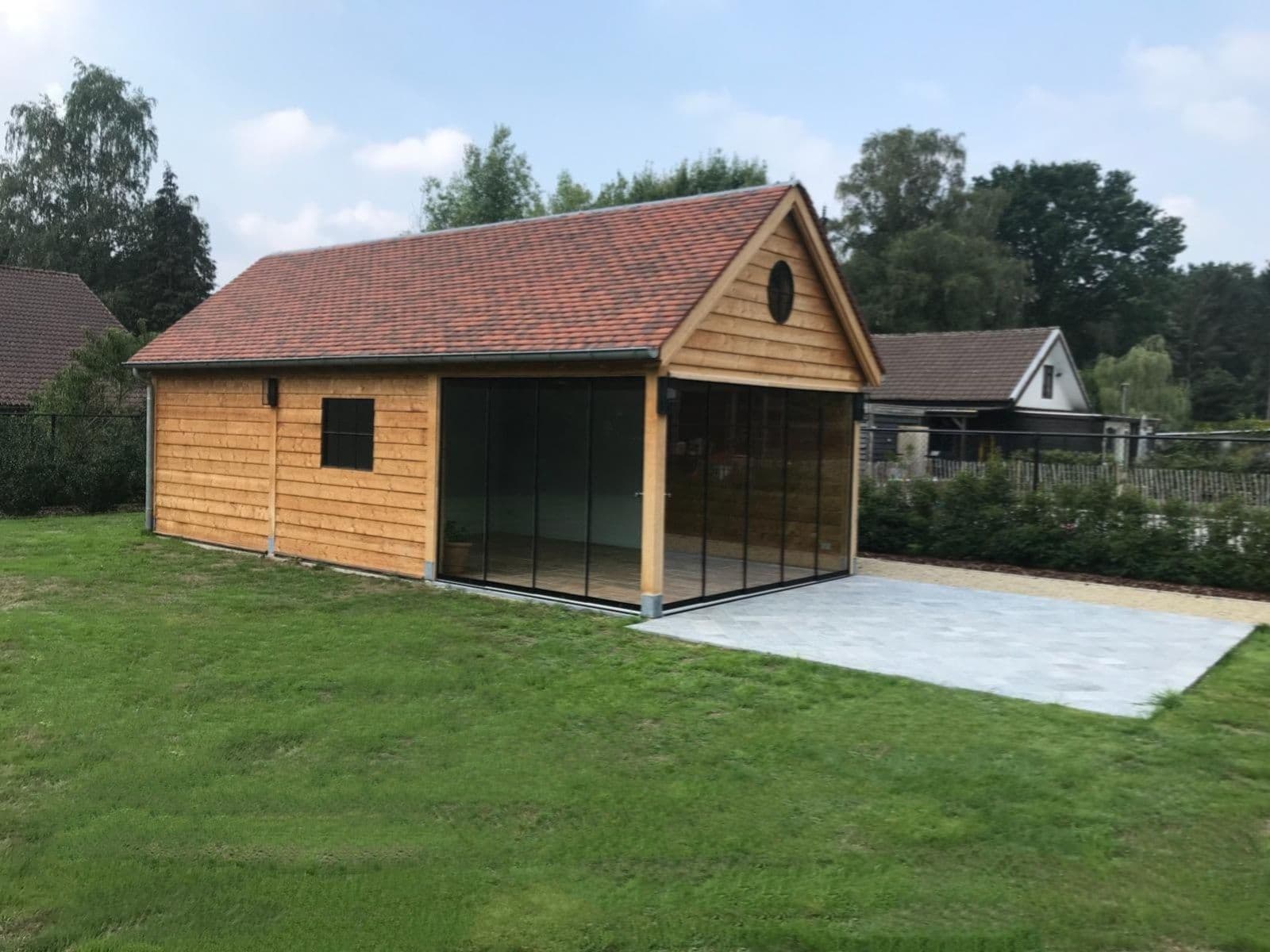 Houten schuur met glazen aanbouw en rood pannendak op een grasveld, met huizen op de achtergrond.
