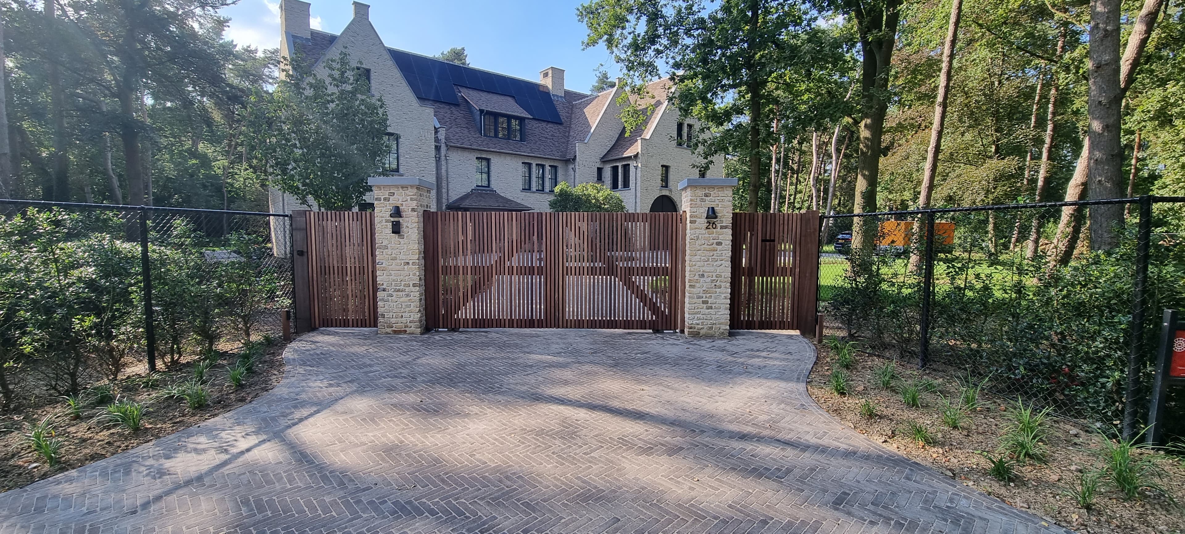 Grote houten dubbele poort tussen stenen pijlers voor een stenen huis met zonnepanelen; bomen en hek eromheen.