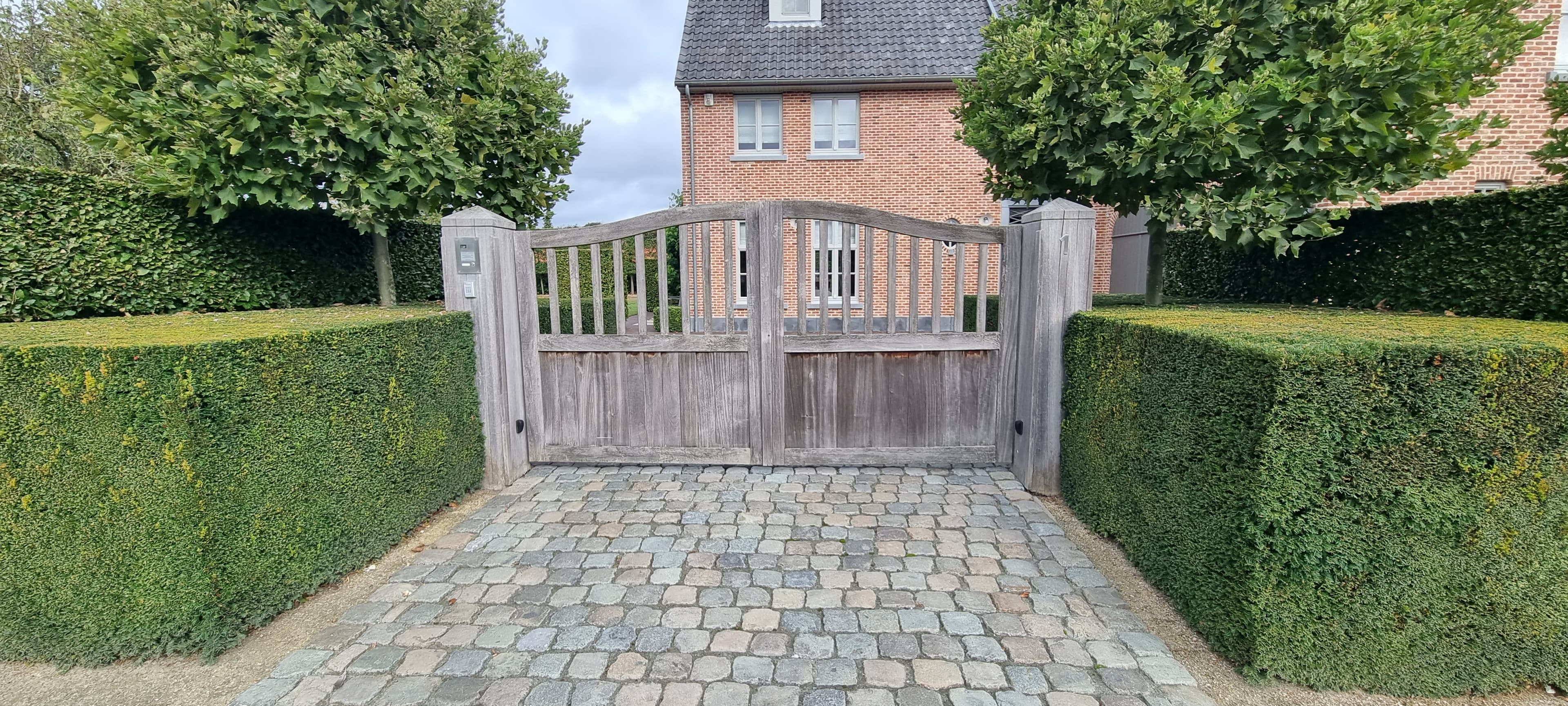 Brede houten poort met verticale spijlen en gebogen bovenkant, tussen groene hagen op een kasseien oprit bij een bakstenen huis.