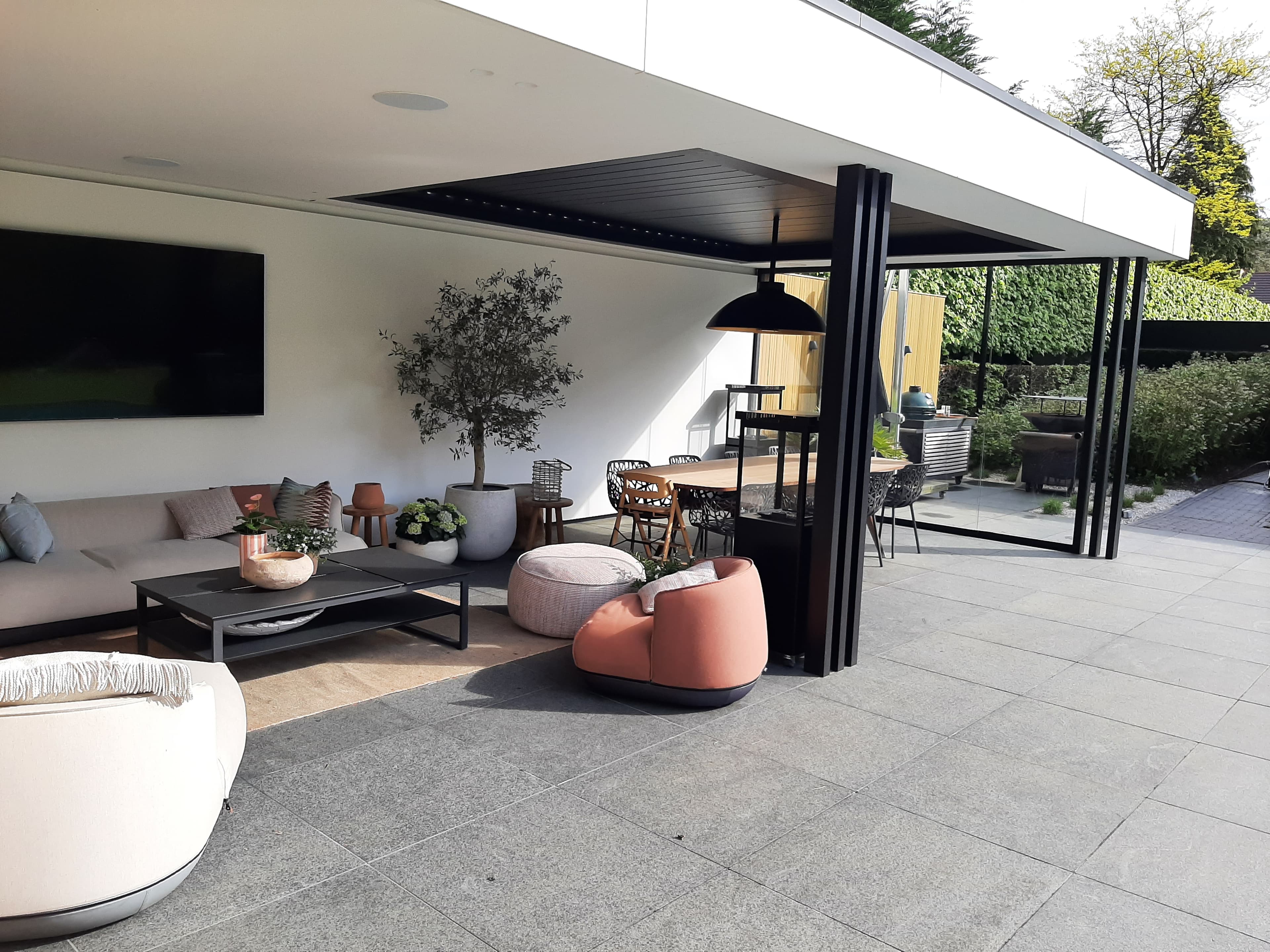 Moderne buitenruimte onder een overdekt terras met lange zithoek, tv aan witte muur, planten en lange eettafel met stoelen.