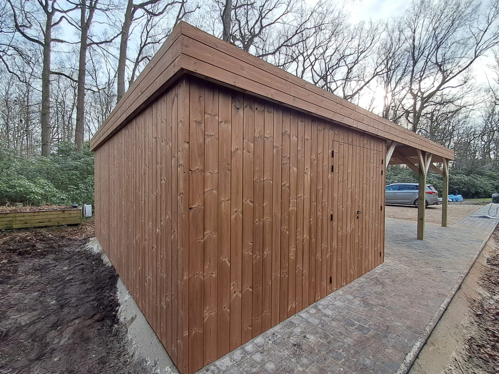 Houten opslagschuur met verticale planken en deur, onder een afdak langs een stenen pad, omgeven door bomen.