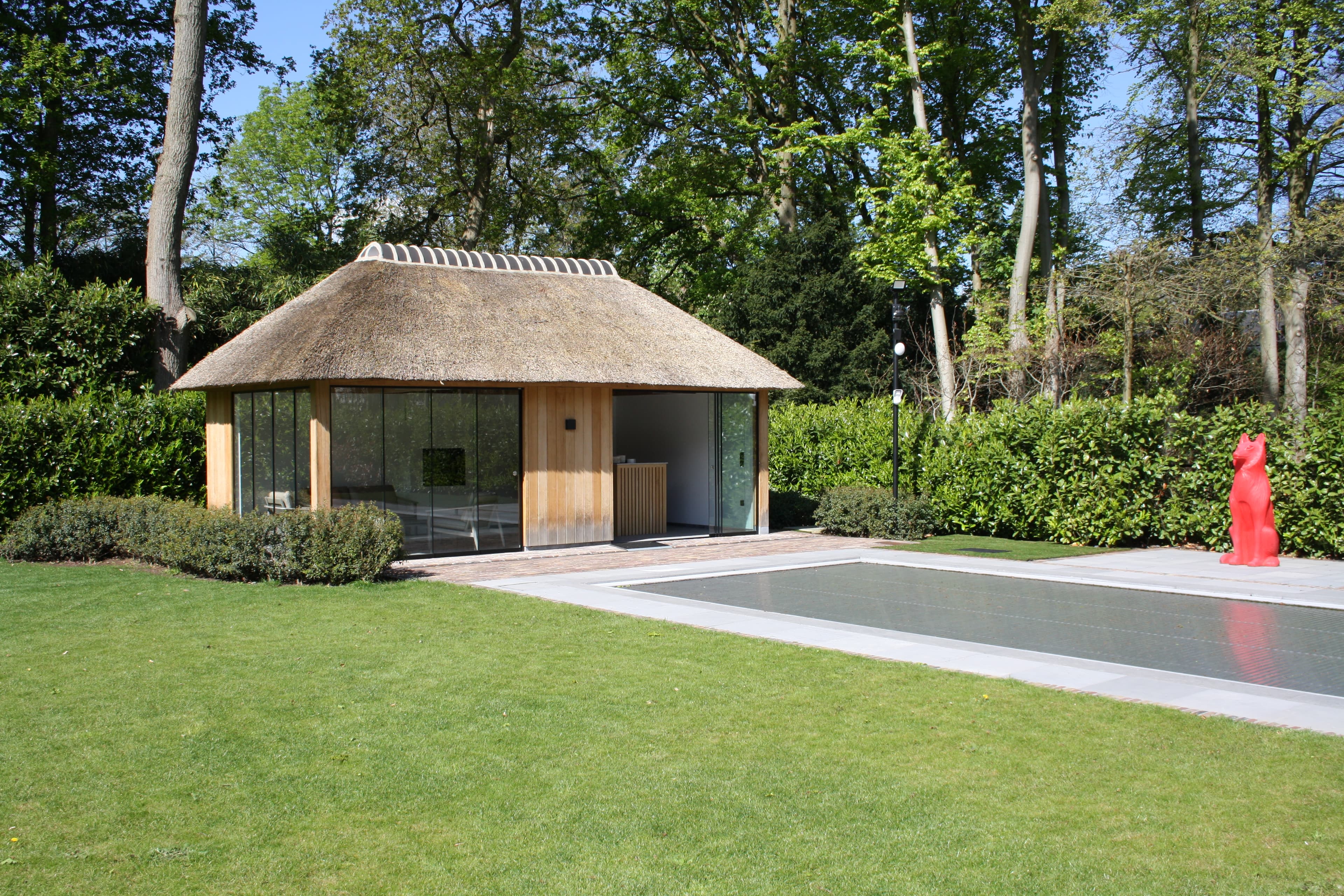Kleine houten hut met rieten dak in een groene tuin, glazen wanden en open deur; rechts een rood standbeeld bij een langwerpige waterpartij.