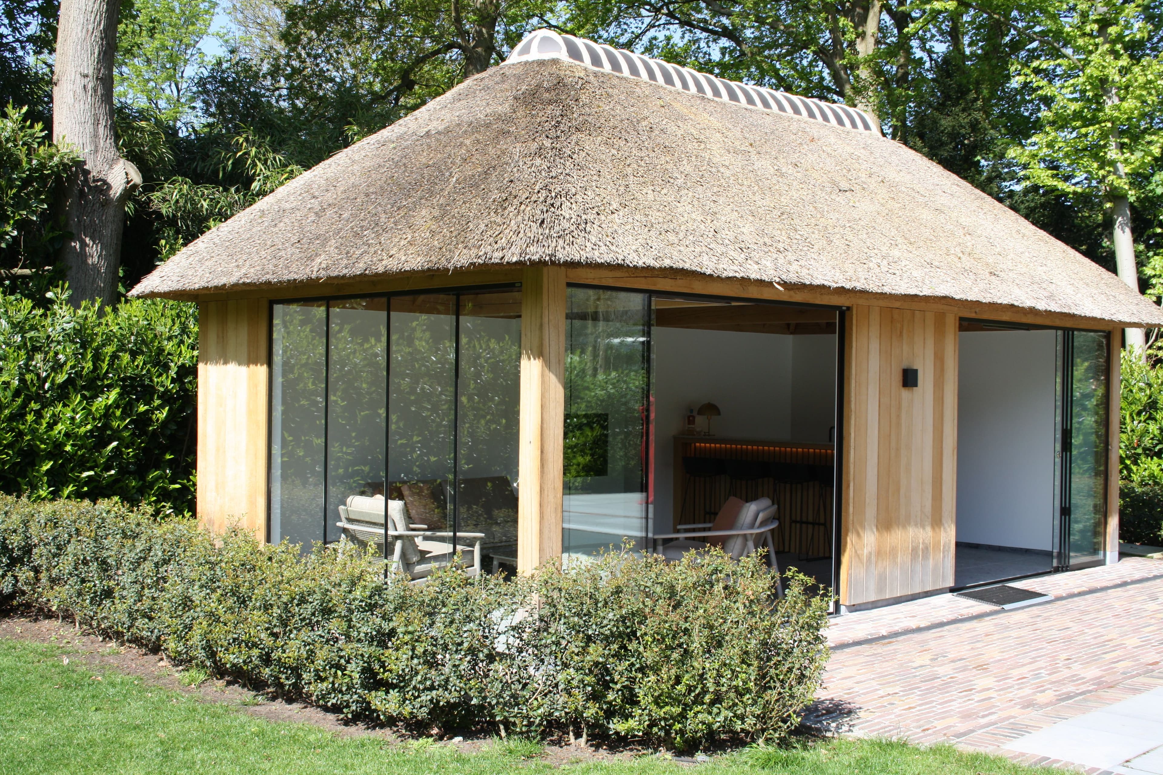 Kleine ronde hut met rieten dak en glazen wanden in een tuin; houten frame en zithoek.