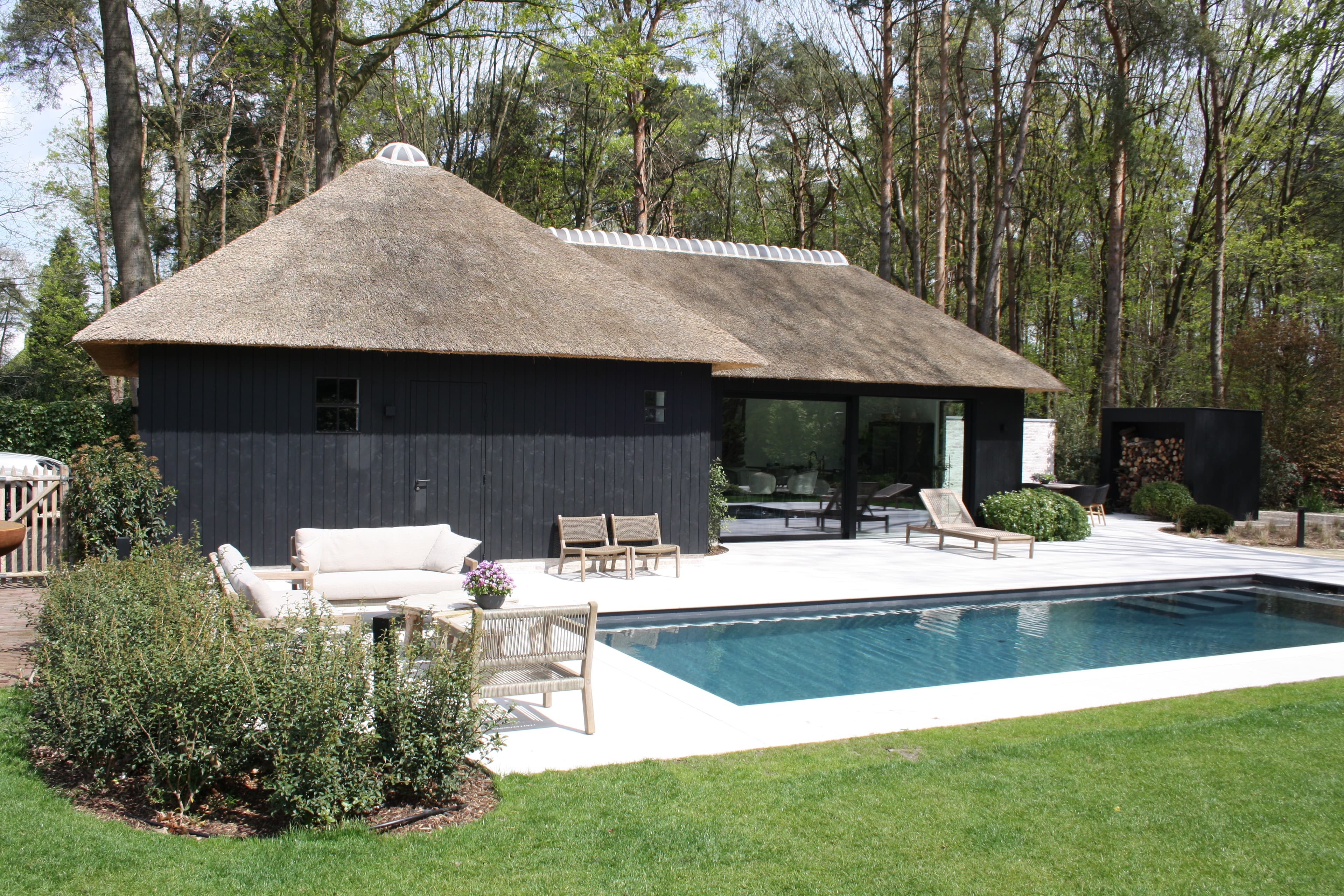 Woonhuis met rieten dak en donkerblauwe houten wanden, glaswand, zwembad en lounge op het terras, omgeven door bomen.