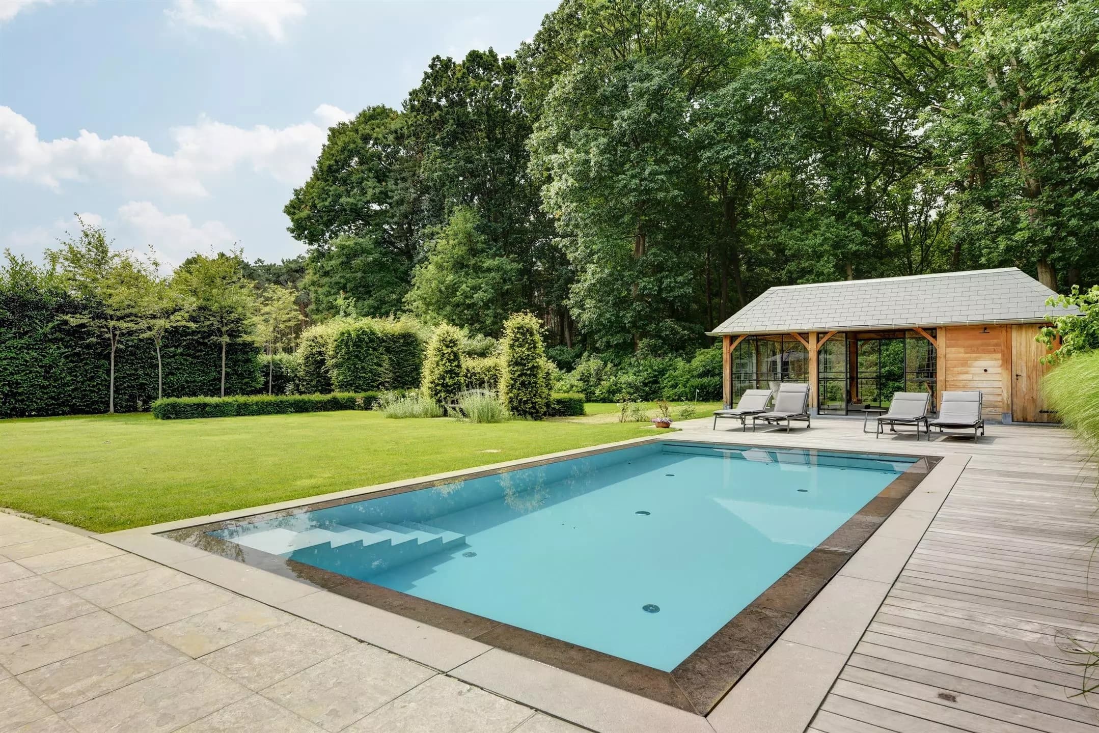 Ruime tuin met helder blauw zwembad, houten poolhouse, ligstoelen en groen gazon omgeven door bomen.