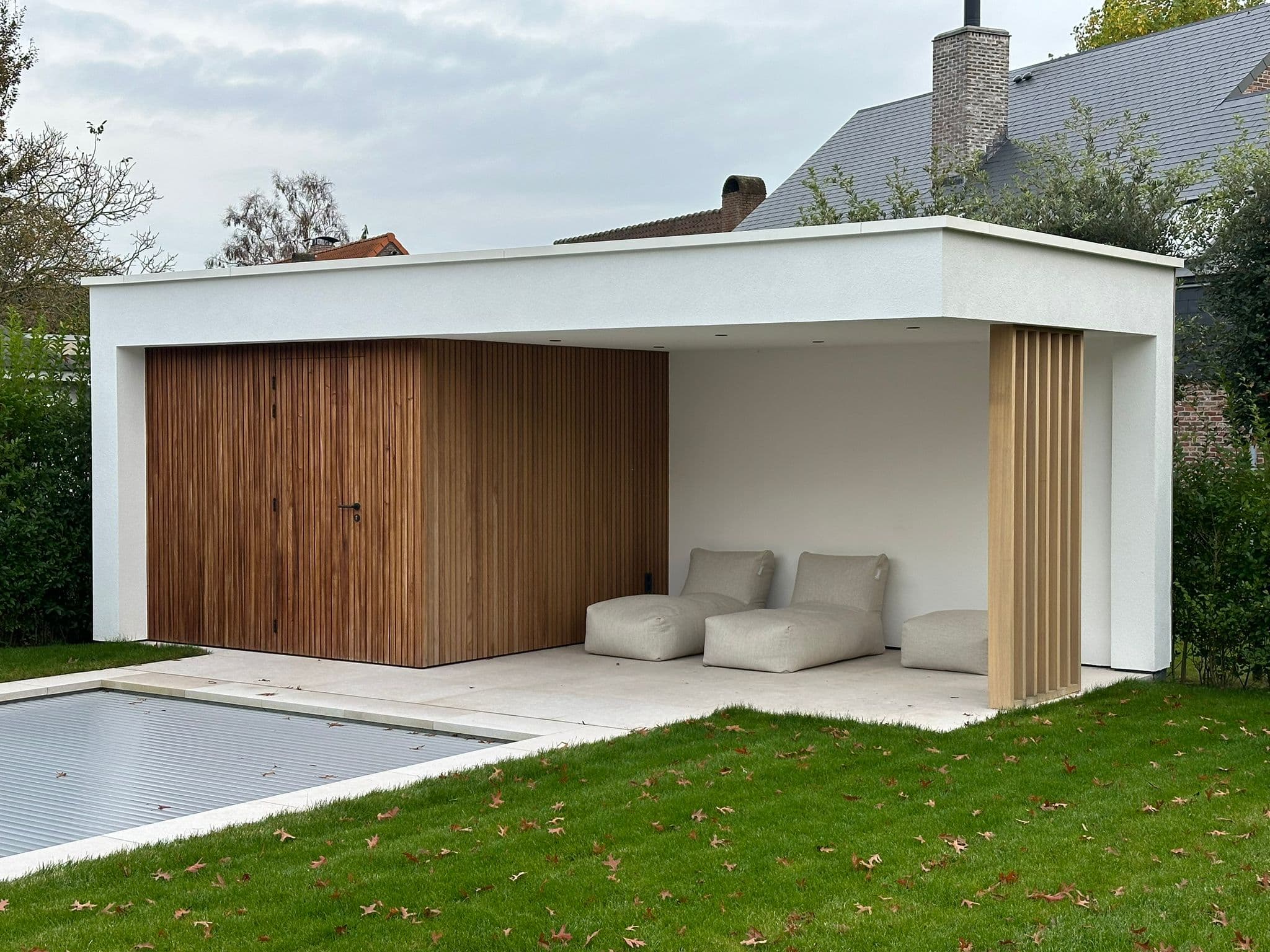 Moderne witte buitenruimte met houten wand en verticale latten, drie beige zitkussens; naast een afgedekt zwembad.