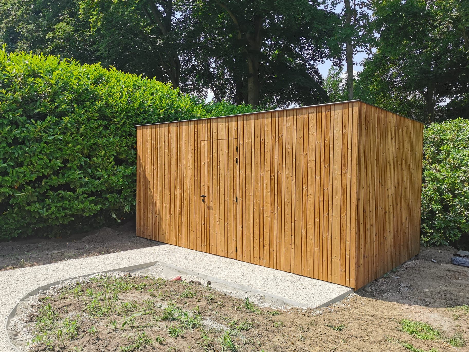 Een houten tuinschuur met verticale planken in een groene tuin met hagen en bomen, op een grindpad.