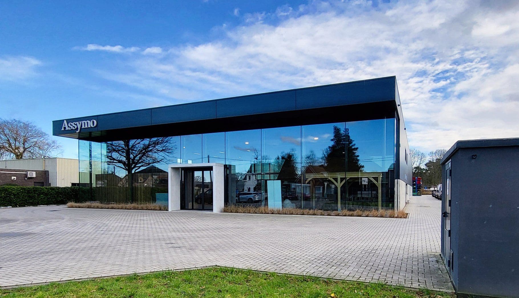 Moderne glazen kantoorgebouw met grote glasgevels en reflecties, logo Assymo aan de linkerkant van het gebouw.