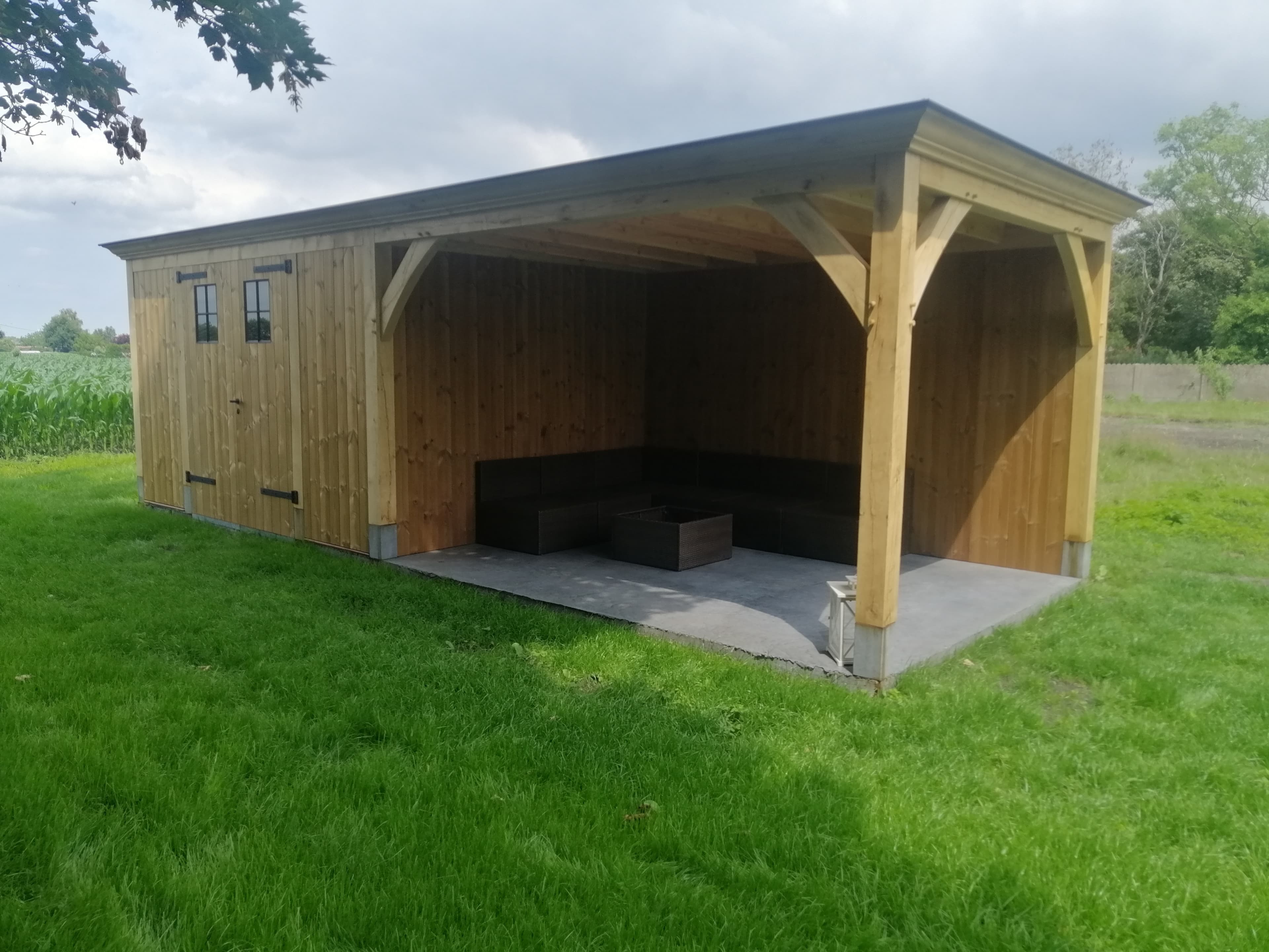 Houten schuur met open voorkant, L-vormige bank en koffietafel op betonnen vloer, groene tuin eromheen.