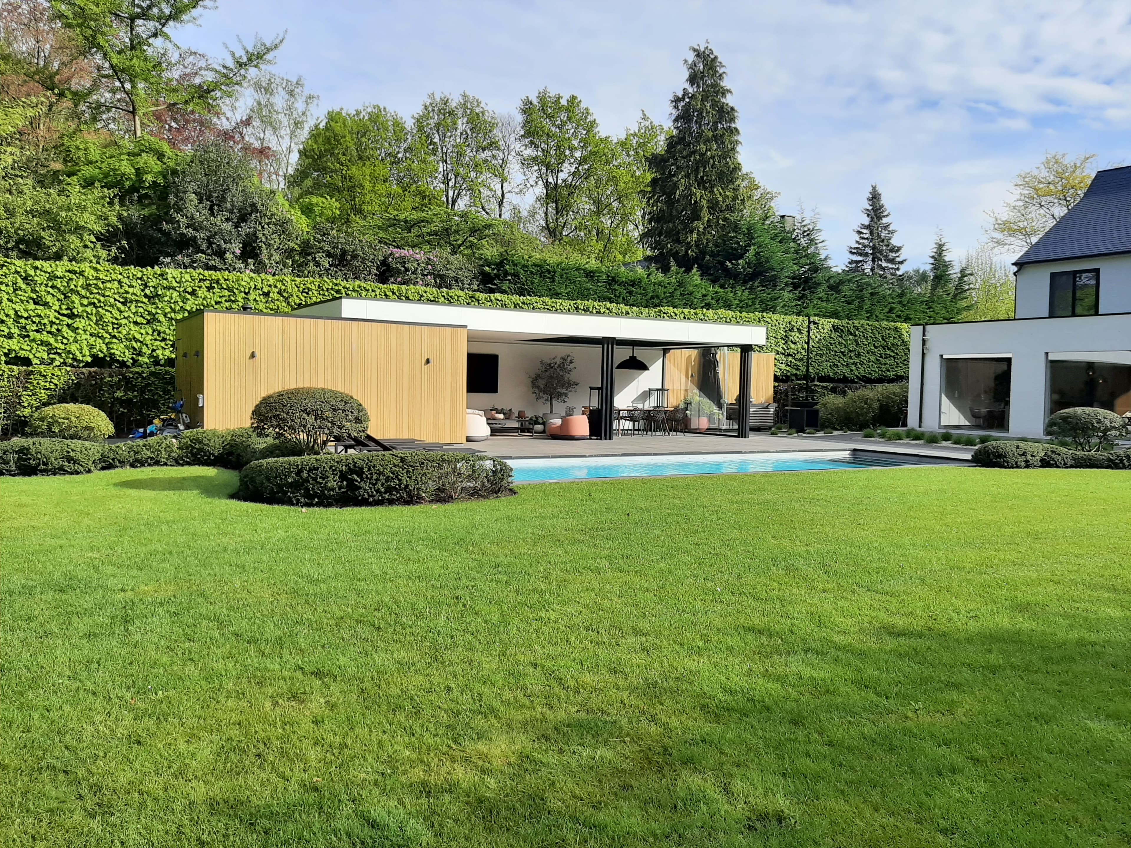 Moderne achtertuin met zwembad, houten paviljoen en buitenmeubilair, omgeven door strak gazon en groene heggen.
