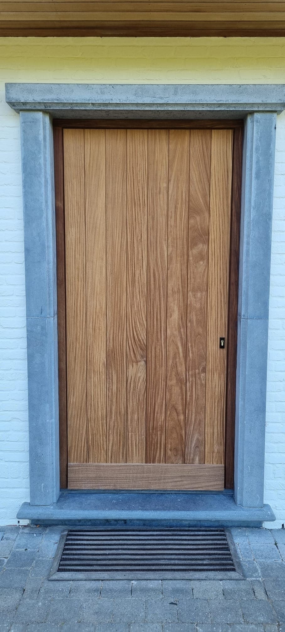 Houten deur met verticale panelen in een grijs stenen omlijsting aan een gele gevel, met een metalen rooster in de stoep voor de deur.