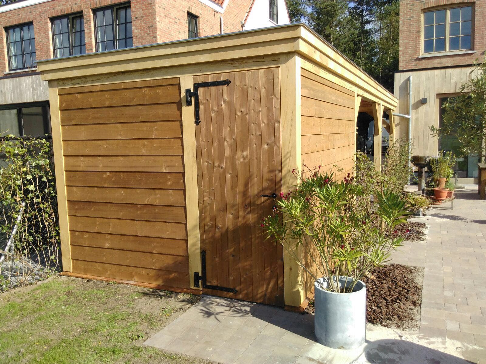 Houten tuinschuur met deur en zwarte scharnieren, langs een betegelde patio met plantenpotten en nabij gebouwen.