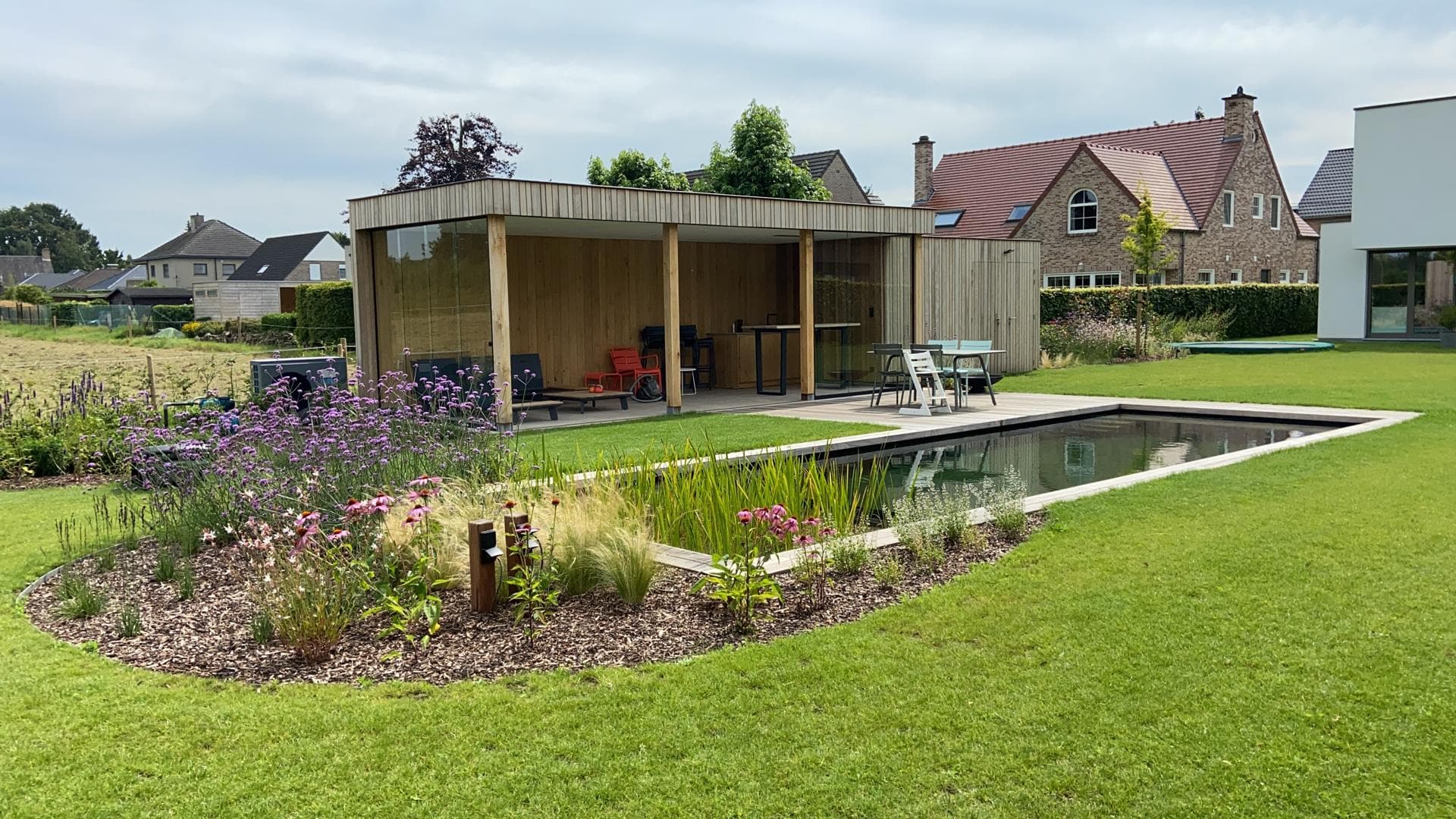 Moderne houten paviljoen met glazen wanden en zitjes langs een lang zwembad, omgeven door een groene tuin met bloemen.