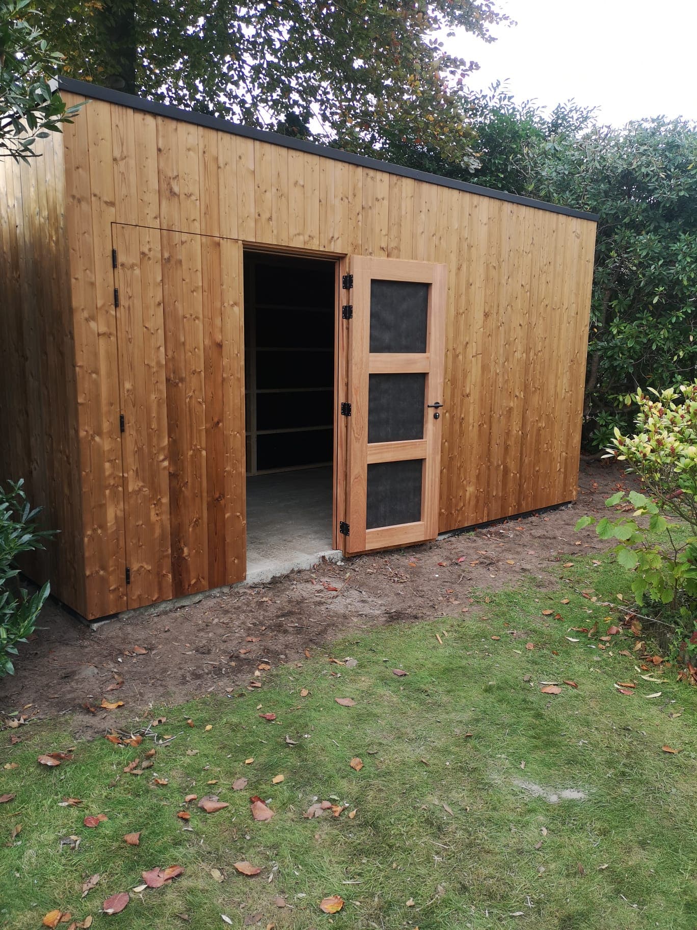 Houten tuinhuisje met open deur; binnen zijn planken zichtbaar, in een tuin met bomen en struiken.