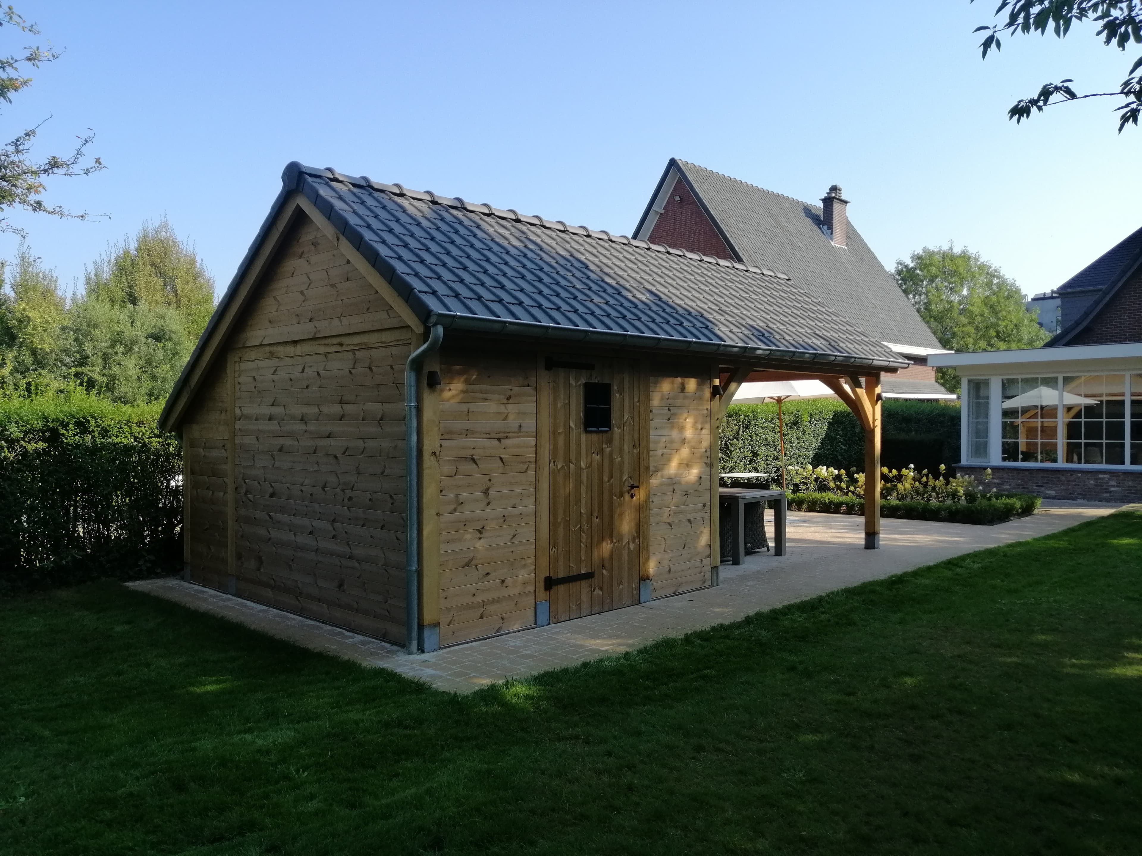 Houten tuinhuis met pannendak op betegelde patio naast een huis met grote ramen; groene tuin met gras en heggen.