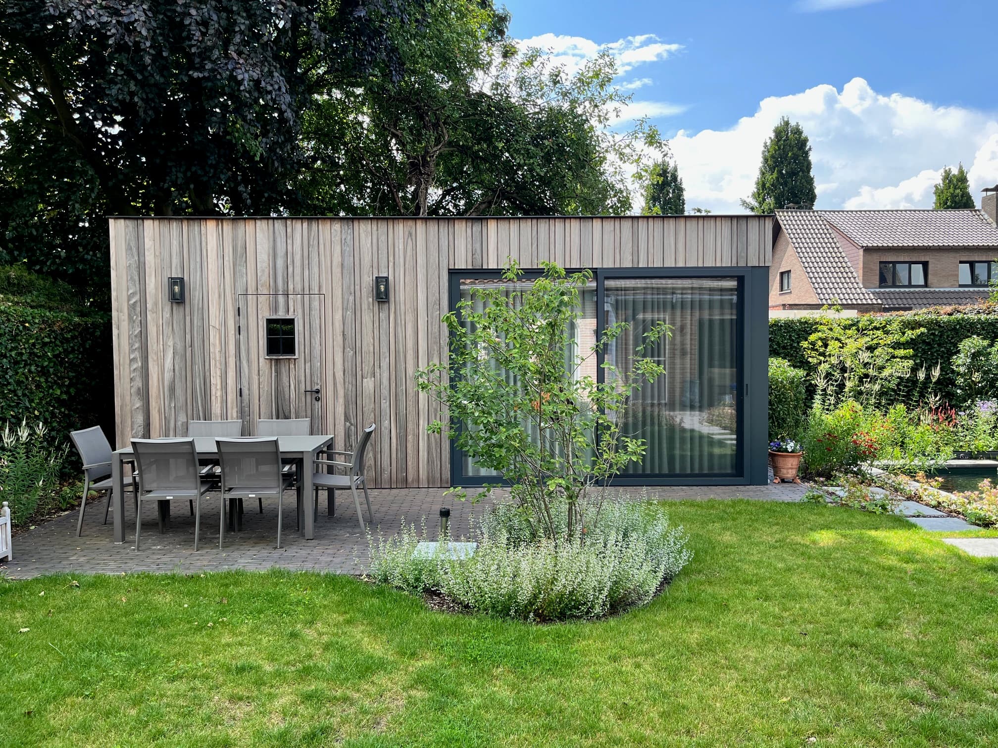 Houten bijgebouw met donkere kozijnen en grote glazen schuifdeur, patio met tafel en stoelen, groene tuin met bomen en bloemen.