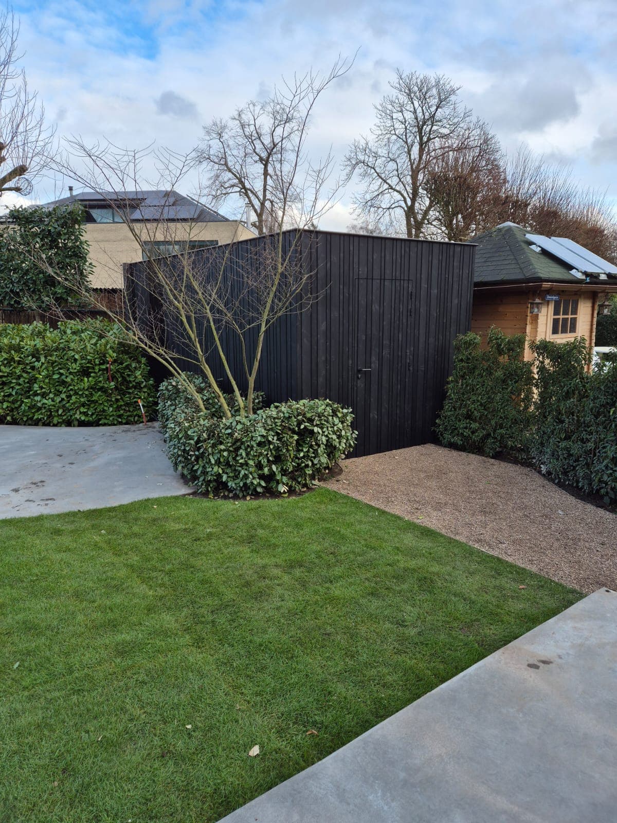Tuin met groen gazon, zwart houten schuurtje, klein houten tuinhuis met zonnepanelen, en groene struiken met kale bomen en een grindpad.